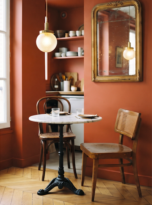 Coin repas vintage dans un petit appartement avec mur terracotta, guéridon en fonte et marbre, chaises dépareillées, suspension boule et miroir ancien.